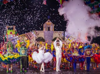 A fotografia exibe uma festa com muitas pessoas usando trajes coloridos e um casal vestido de branco no centro. Há fumaça do lado direito, e confetes do lado esquerdo. O céu está escuro e no fundo há uma igreja.