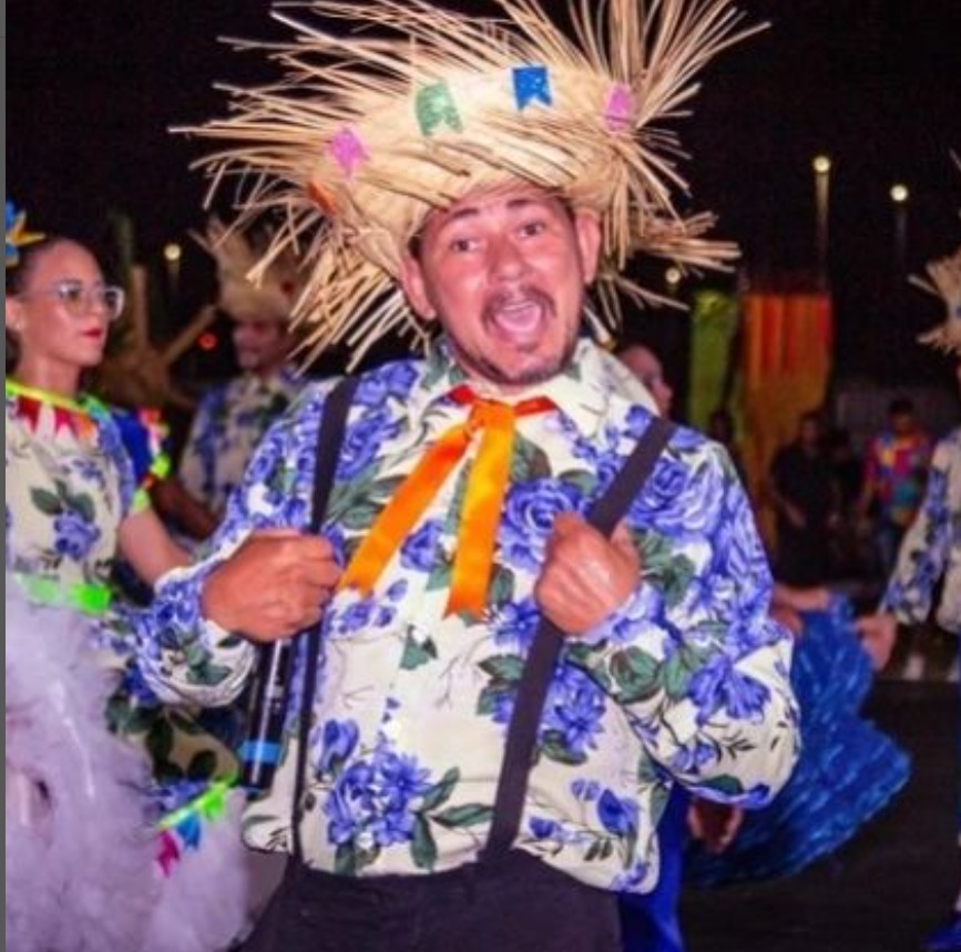 Homem com chapéu de palha, camisa branca com flores azuis, fita laranja no colarinho e suspensório preto.